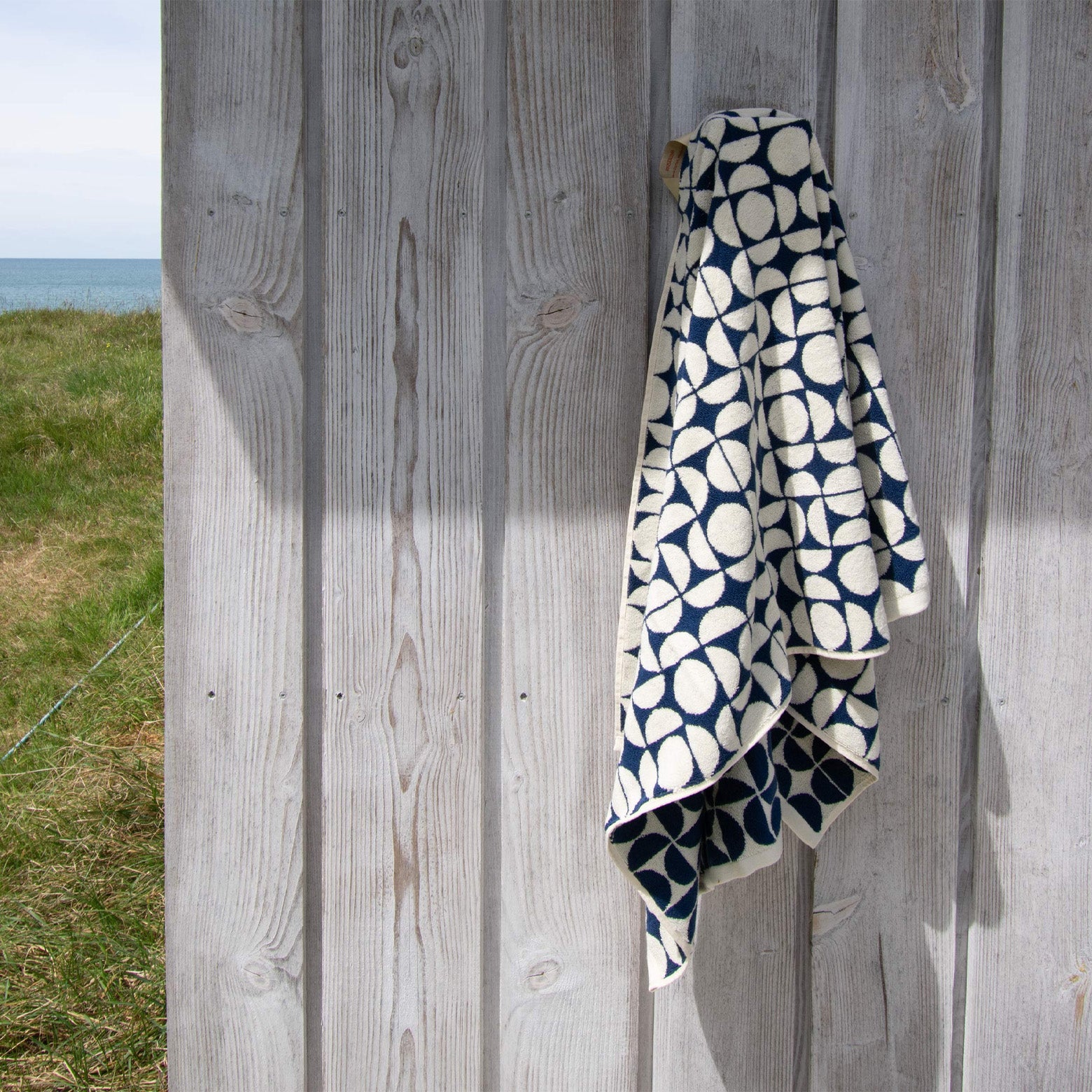 Badehåndklæde med blå og hvid grafisk mønster stylet ved strand. Homehagen. 140 x 70 cm
