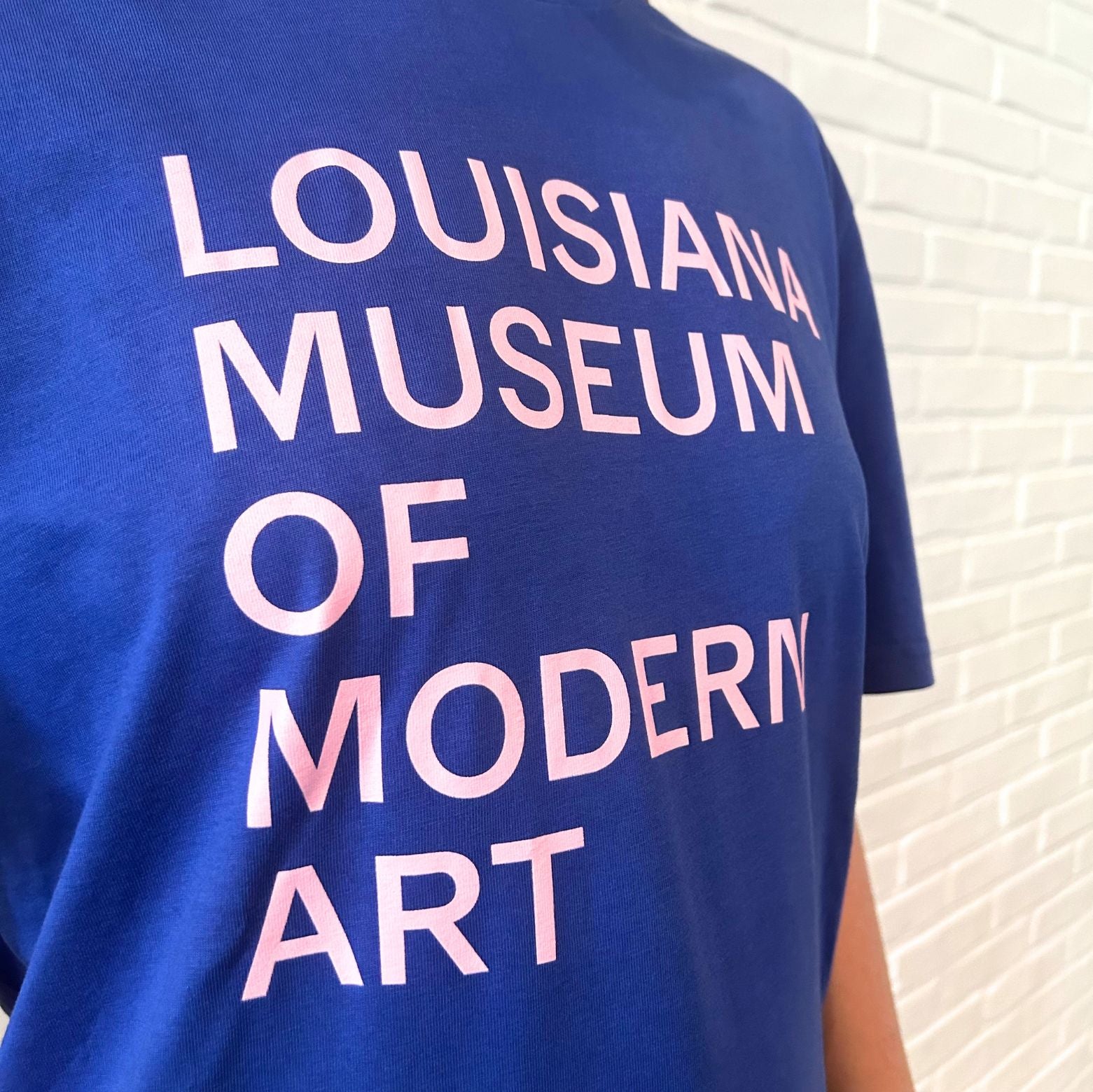 Louisiana T-shirt i blå med teksten “Louisiana Museum of Modern Art” i lyserødt print stylet i Louisianas interiør. 